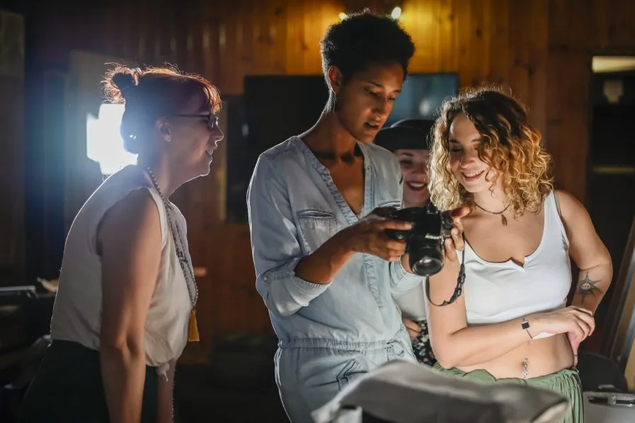 L'image montre trois personnes rassemblées autour d'un appareil photo. Une personne tient l'appareil photo tandis que les deux autres le regardent attentivement. Le cadre semble être en intérieur, avec des murs en bois et une source de lumière en arrière-plan. Les individus semblent passer en revue ou discuter de quelque chose lié à la photographie ou à la vidéographie. Cette scène capture un moment de collaboration et d'apprentissage, mettant en avant l'interaction et le focus sur l'appareil photo. Il n'y a aucun problème de mathématiques ou de devoir visible sur l'image.